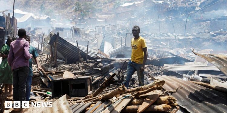 Man in yellow t-shirt stands in piles of rubble and corrugated iron while three other people stand on the left of the picture. The rubble seems to go on for some distance and the air is hazy.
