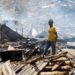 Man in yellow t-shirt stands in piles of rubble and corrugated iron while three other people stand on the left of the picture. The rubble seems to go on for some distance and the air is hazy.
