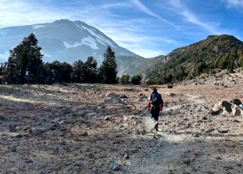 A rescuer hikes along rough terrain and rocks toward Mount Shasta
