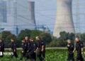 About a dozen German police officers in black overalls are seen combing a field during a search. An active power station is seen in the background.