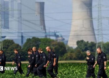 About a dozen German police officers in black overalls are seen combing a field during a search. An active power station is seen in the background.