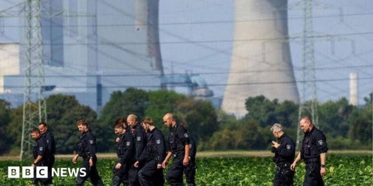 About a dozen German police officers in black overalls are seen combing a field during a search. An active power station is seen in the background.