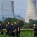 About a dozen German police officers in black overalls are seen combing a field during a search. An active power station is seen in the background.