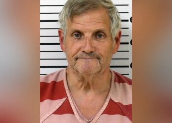 Booking photo of John Herb Hayes III, a gray-haired man wearing a red-and-pink striped shirt in front of a height chart.