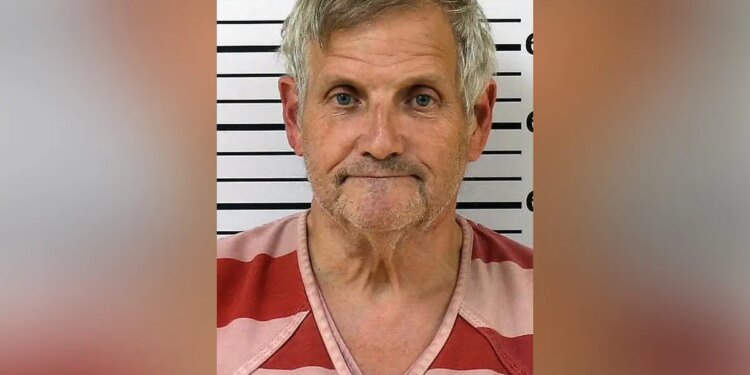 Booking photo of John Herb Hayes III, a gray-haired man wearing a red-and-pink striped shirt in front of a height chart.