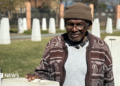 Abraham Mofokeng in a brown patterned cardigan and beanie hat stands by a white monolith. Out of focus, in the background, are other white monoliths - they are part of a memorial.