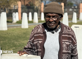 Abraham Mofokeng in a brown patterned cardigan and beanie hat stands by a white monolith. Out of focus, in the background, are other white monoliths - they are part of a memorial.