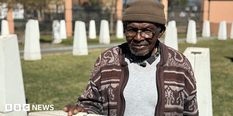Abraham Mofokeng in a brown patterned cardigan and beanie hat stands by a white monolith. Out of focus, in the background, are other white monoliths - they are part of a memorial.