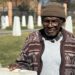 Abraham Mofokeng in a brown patterned cardigan and beanie hat stands by a white monolith. Out of focus, in the background, are other white monoliths - they are part of a memorial.