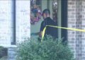 An investigator wearing sunglasses and gloves stands behind yellow police tape outside a brick home.