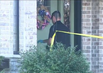 An investigator wearing sunglasses and gloves stands behind yellow police tape outside a brick home.