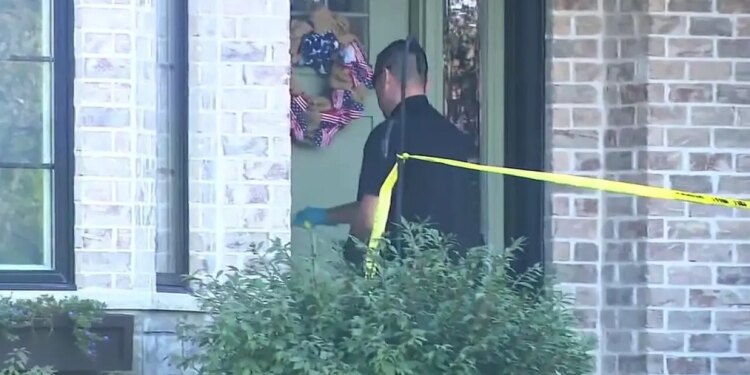 An investigator wearing sunglasses and gloves stands behind yellow police tape outside a brick home.