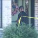 An investigator wearing sunglasses and gloves stands behind yellow police tape outside a brick home.