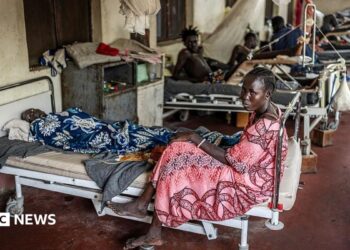 A woman sits at the foot of a bed that has a male patient on it. There are filled beds behind her.