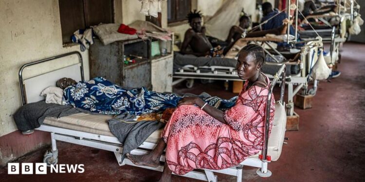A woman sits at the foot of a bed that has a male patient on it. There are filled beds behind her.