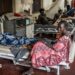 A woman sits at the foot of a bed that has a male patient on it. There are filled beds behind her.