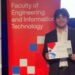 A young man in a suit holding up a certificate and standing in front of signs, one titled UTS Tech Festival and another Faculty of Engineering and Information Technology