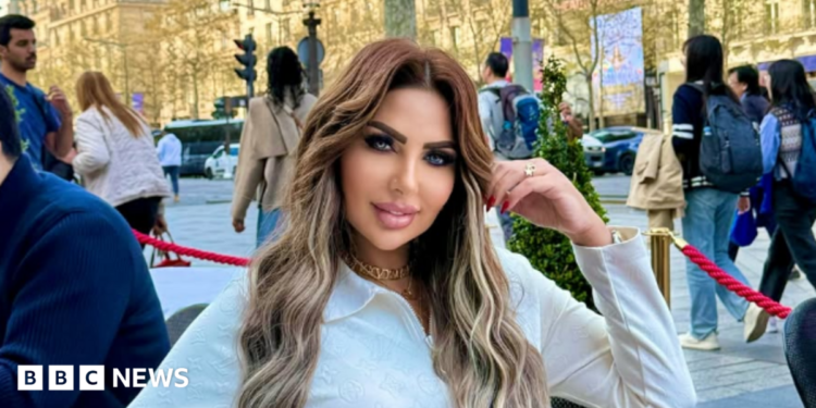 Sarah Khalifa wearing a white long-shirted with her folded left arm, the hand touching her long hair, sits by the side of a road inside a red-roped cordoned area, beyong which people walk on a road the other side of which has rows of sunbathed buildings