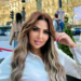 Sarah Khalifa wearing a white long-shirted with her folded left arm, the hand touching her long hair, sits by the side of a road inside a red-roped cordoned area, beyong which people walk on a road the other side of which has rows of sunbathed buildings