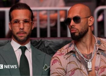 Tristan Tate is wearing sunglasses and is dressed in a green suit and tie and white shirt. Andrew is to his right in sunglasses and an open necked shirt.