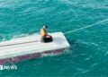 A teenage boy, wearing a rain jacket, sitting on his knees on top of an overturned boat surrounded by blue-green waters.