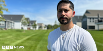 Gerald Bourguet, a white man standing in a field of a manicured lawn. He is wearing a white shirt and crucifix chain. There are homes in the background that are blurred.