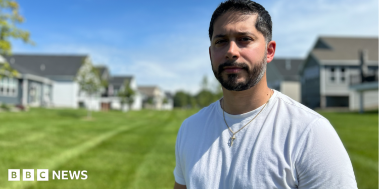 Gerald Bourguet, a white man standing in a field of a manicured lawn. He is wearing a white shirt and crucifix chain. There are homes in the background that are blurred.