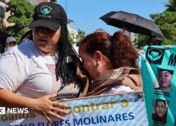 People take part in a march for peace in Culiacan, Mexico on 13 September 2026. A woman wearing a white T-shirt and a black baseball cap comforts another woman who is crying. The woman with the baseball cap is carrying a placard reading "help us find Jose Gonzalo Flores Molinares". In the background another protester is holding up another banner with the photos of missing people on it.