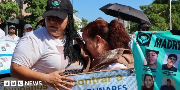 People take part in a march for peace in Culiacan, Mexico on 13 September 2026. A woman wearing a white T-shirt and a black baseball cap comforts another woman who is crying. The woman with the baseball cap is carrying a placard reading "help us find Jose Gonzalo Flores Molinares". In the background another protester is holding up another banner with the photos of missing people on it.