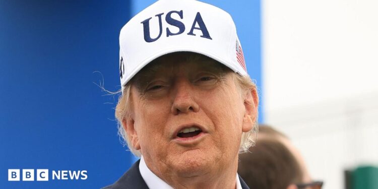 U.S. President Donald Trump attends the Amgen Irish Open at Trump International Golf Links in Doonbeg, Ireland. He is wearing a white USA baseball cap, navy blazer with a white shirt. He has his arms outstretched.