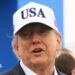U.S. President Donald Trump attends the Amgen Irish Open at Trump International Golf Links in Doonbeg, Ireland. He is wearing a white USA baseball cap, navy blazer with a white shirt. He has his arms outstretched.