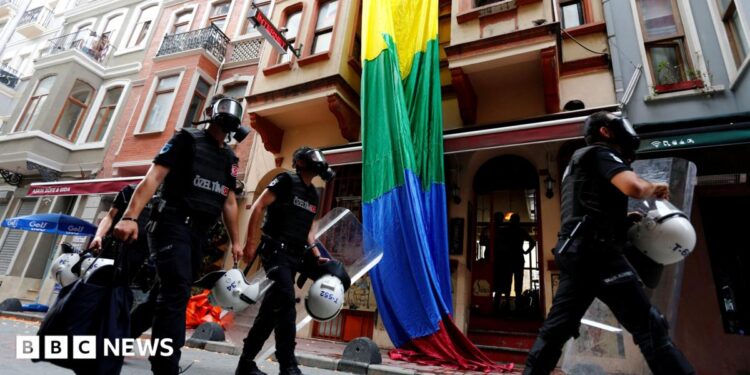 Riot police, with a rainbow flag in the background, chase LGBT activists as they try to gather for a pride parade, which was banned by the governorship in Istanbul. Officers are wearing black uniforms, wearing gas masks, and are carrying helmets and riot shields as they move through what appears to be a residential street.