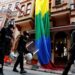 Riot police, with a rainbow flag in the background, chase LGBT activists as they try to gather for a pride parade, which was banned by the governorship in Istanbul. Officers are wearing black uniforms, wearing gas masks, and are carrying helmets and riot shields as they move through what appears to be a residential street.