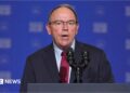 Close-up of Tom Tiffany, wearing a suit and tie, speaking into a microphone at a podium in front of a blue background