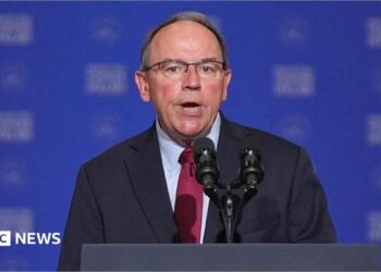 Close-up of Tom Tiffany, wearing a suit and tie, speaking into a microphone at a podium in front of a blue background