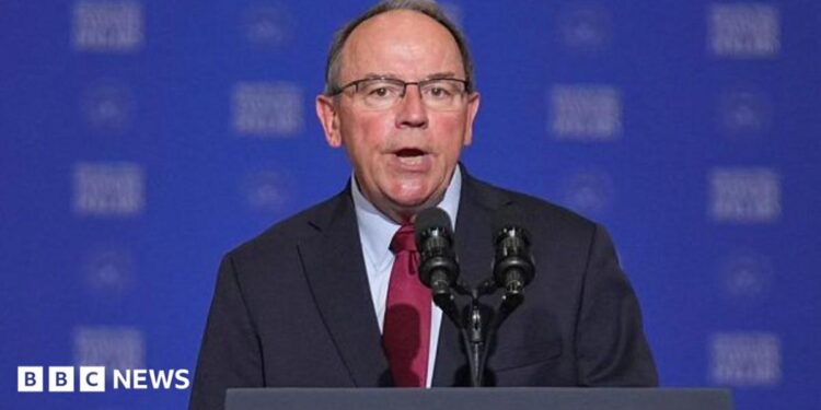 Close-up of Tom Tiffany, wearing a suit and tie, speaking into a microphone at a podium in front of a blue background