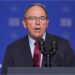 Close-up of Tom Tiffany, wearing a suit and tie, speaking into a microphone at a podium in front of a blue background