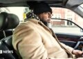 A bearded man in a beige coat and a black and white chequered scarf drives his car through a US urban area. His car has a black leather interior.