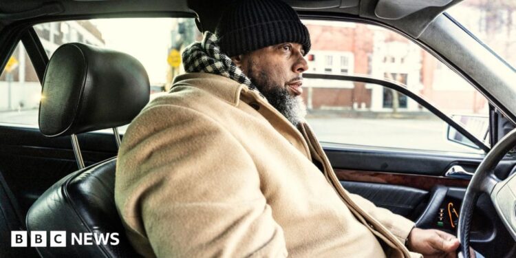 A bearded man in a beige coat and a black and white chequered scarf drives his car through a US urban area. His car has a black leather interior.
