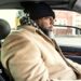 A bearded man in a beige coat and a black and white chequered scarf drives his car through a US urban area. His car has a black leather interior.