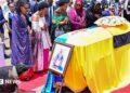 Family members and mourners attend a ceremony to receive the coffin with the body of King Oyo Nyimba Kabamba Iguru Rukidi IV at Entebbe airport. The casket is draped in the Tooro kingdom flag and the Ugandan flag. A portrait of the monarch has been place at the front of the coffin and woman in a black shawl is kneeling before it.