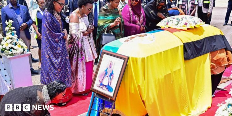 Family members and mourners attend a ceremony to receive the coffin with the body of King Oyo Nyimba Kabamba Iguru Rukidi IV at Entebbe airport. The casket is draped in the Tooro kingdom flag and the Ugandan flag. A portrait of the monarch has been place at the front of the coffin and woman in a black shawl is kneeling before it.