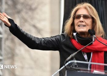 Gloria Steinem, in a red scarf and sunglasses, gestures with an outstretched arm as she speaks at a podium