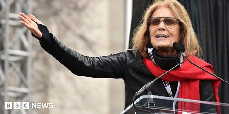 Gloria Steinem, in a red scarf and sunglasses, gestures with an outstretched arm as she speaks at a podium