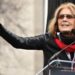 Gloria Steinem, in a red scarf and sunglasses, gestures with an outstretched arm as she speaks at a podium