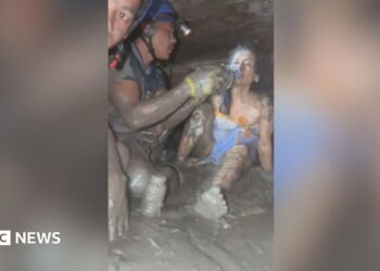 A woman lying in mud being fed water from a bottle by rescuers.
