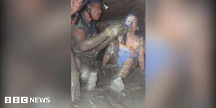 A woman lying in mud being fed water from a bottle by rescuers.