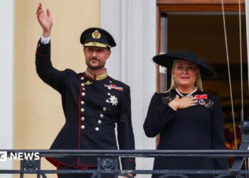 King Haakon waves to the crowd from the balcony of the royal palace, as Queen Mette-Marit stands next to him