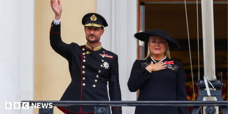 King Haakon waves to the crowd from the balcony of the royal palace, as Queen Mette-Marit stands next to him