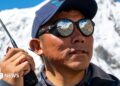 A Nepalese man wearing a cap and sunglasses, holding a walkie-talkie with snowy mountains behind him